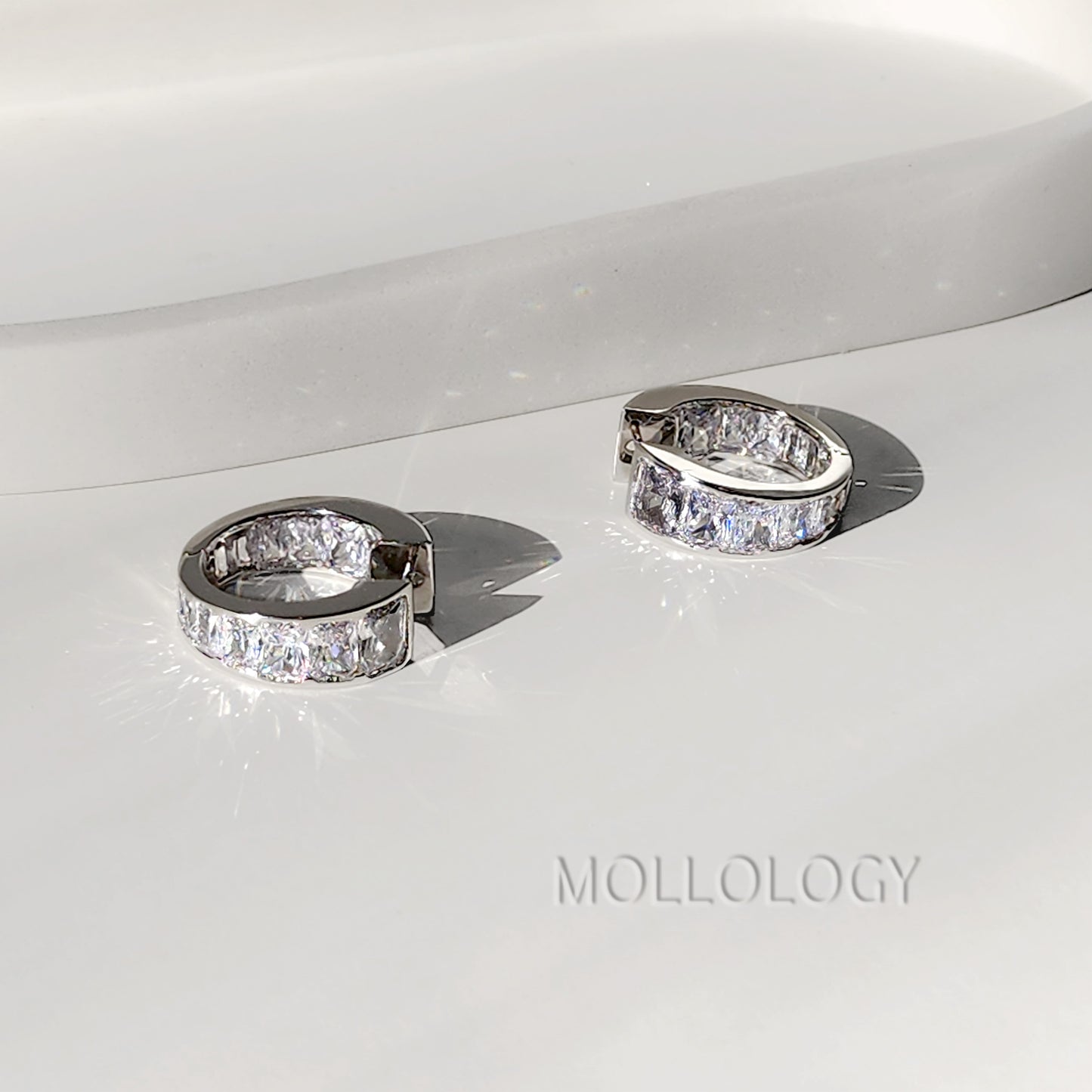 A pair of silver-toned huggie hoop earrings with clear pavé-set stones, displayed on a glossy white surface with a soft shadow. The brand name 'MOLLOLOGY' is printed in a faint gray font in the lower right corner.