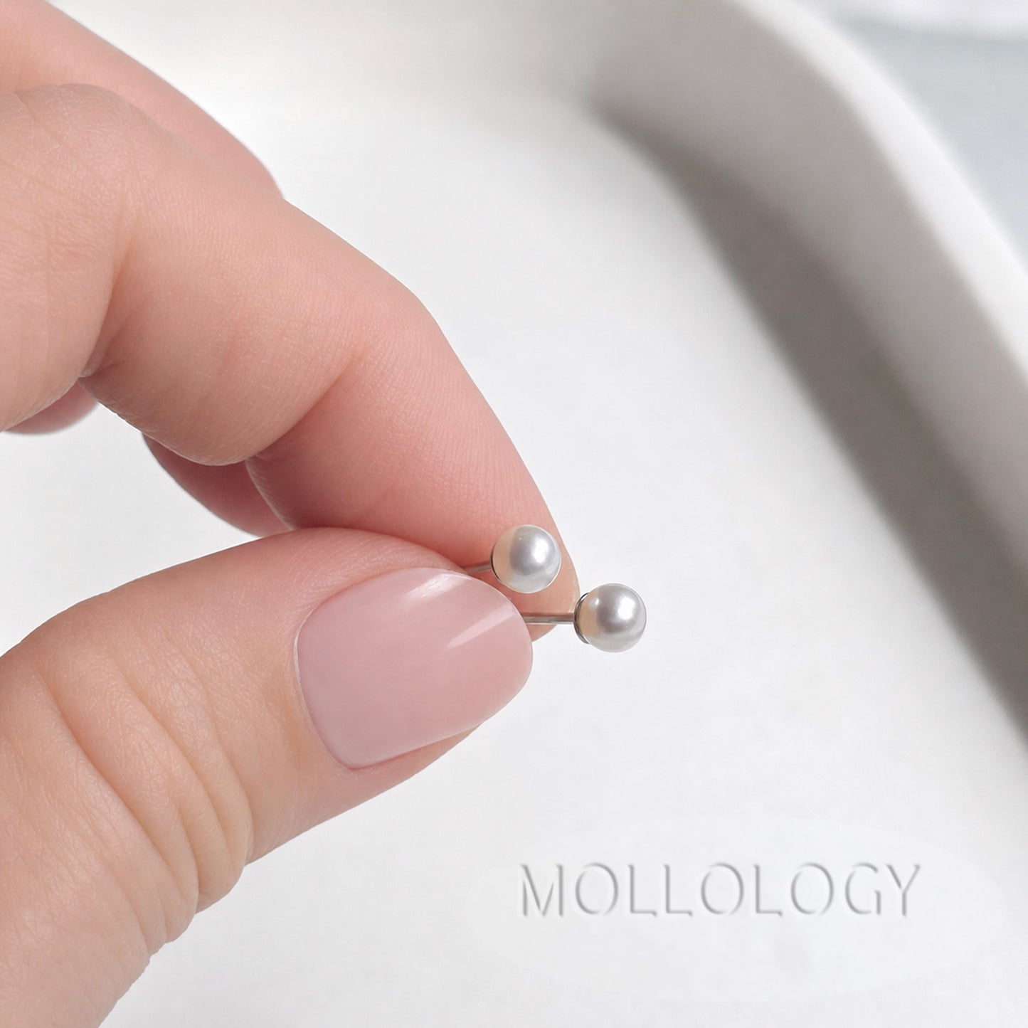 A close-up of a hand with pale pink manicured nails holding a pair of silver-toned pearl stud earrings with small white pearls, against a soft white background with the brand name MOLLOLOGY visible.