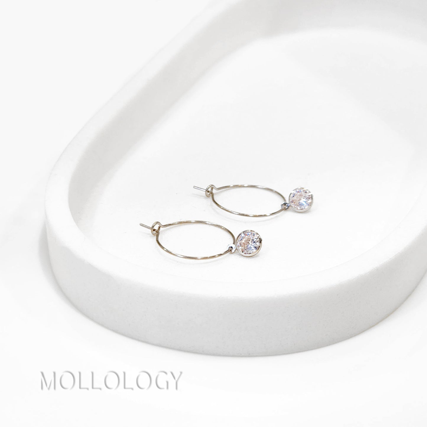 Pair of silver hoop earrings with clear gemstones on a white surface, branded 'Molloy'.
