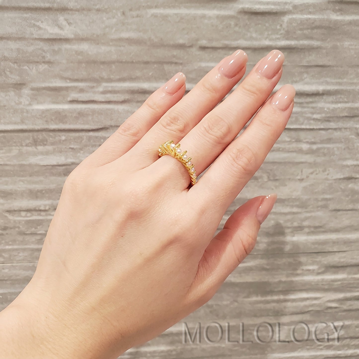 Dainty gold pearl ring on manicured hand with nude nails against ribbed stone background, minimalist stacking jewelry