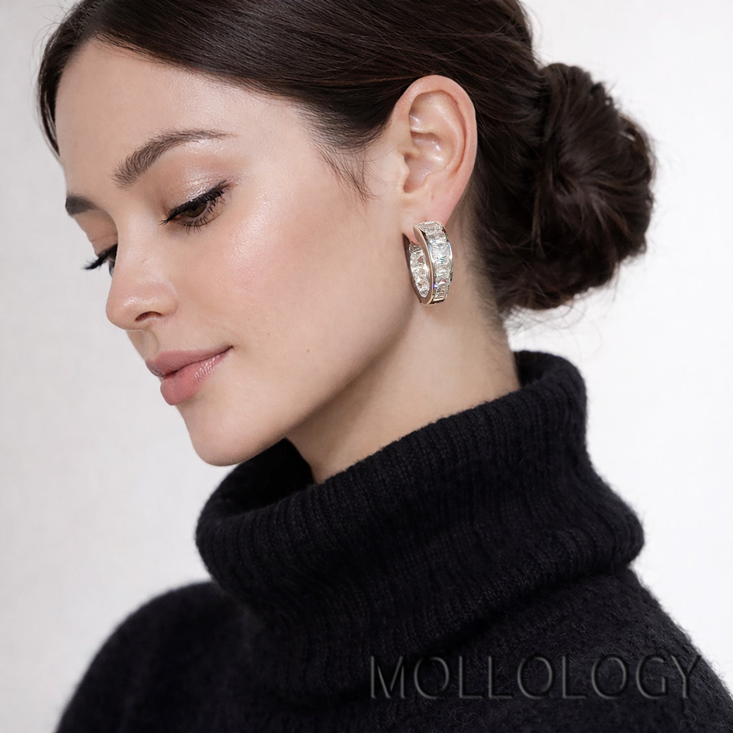 A close-up of a woman wearing a silver hoop earring adorned with clear crystals, paired with a black turtleneck sweater. The brand name MOLLOLOGY is visible as a watermark at the bottom.