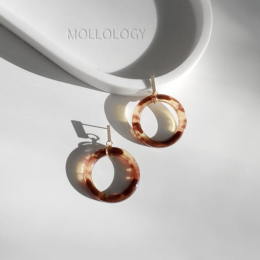 Tortoiseshell acrylic circle dangle earrings with gold bar stud post, flat lay on white ceramic tray with soft natural light and warm shadows