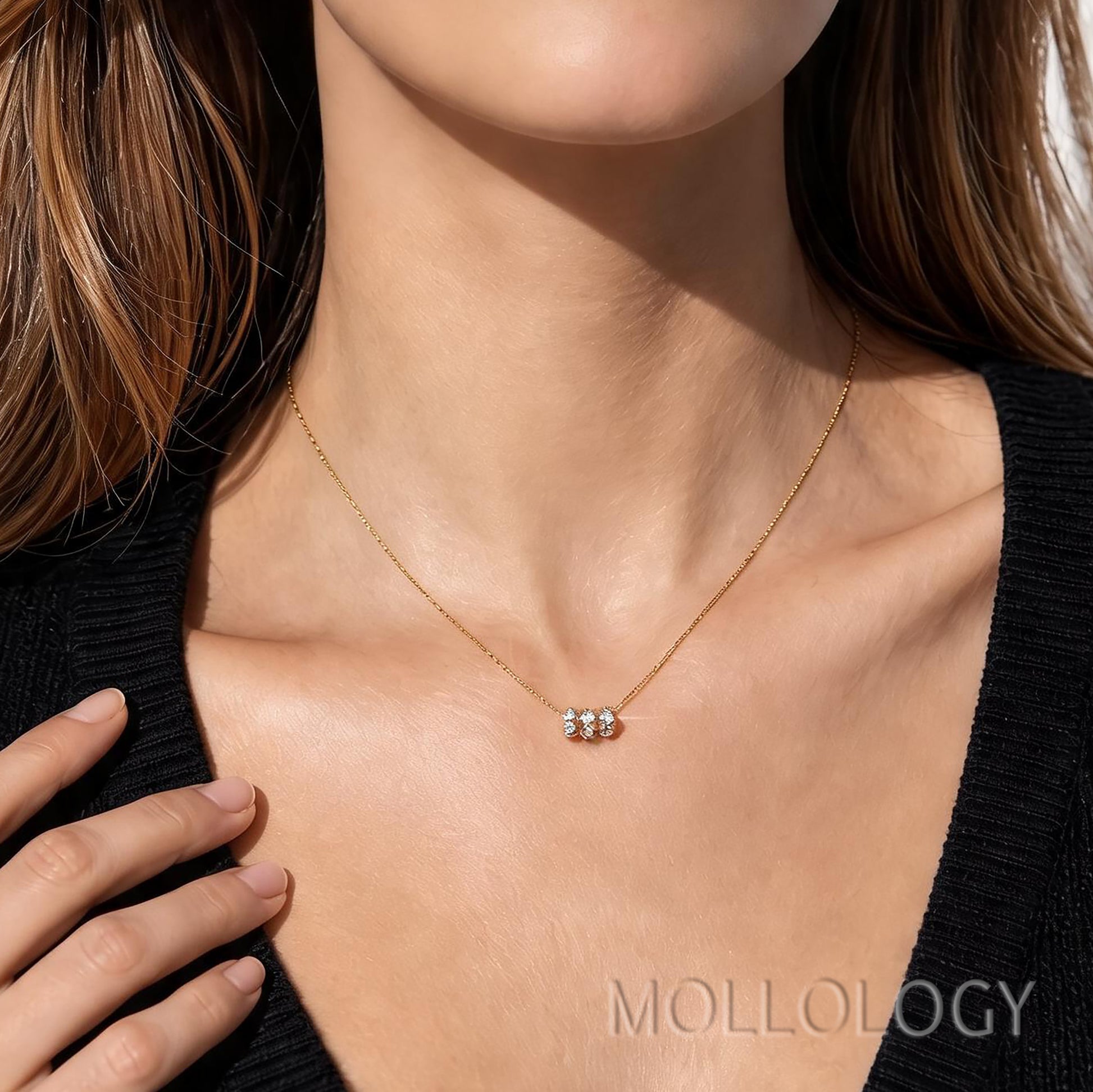 A close-up of a model wearing a delicate gold chain necklace with a small bow-shaped charm set with clear stones. The model is wearing a black knit top, and the brand name 'MOLLOLOGY' is visible as a watermark at the bottom right.