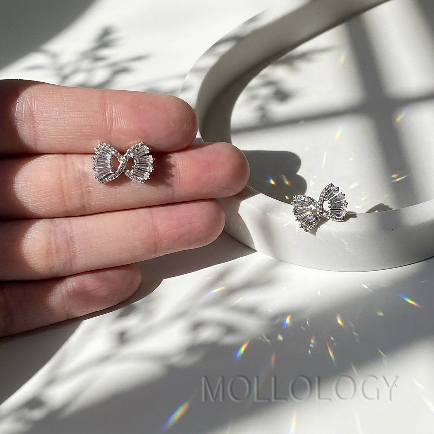 Pair of silver baguette bow stud earrings on white tray, minimalist crystal studs casting soft shadows