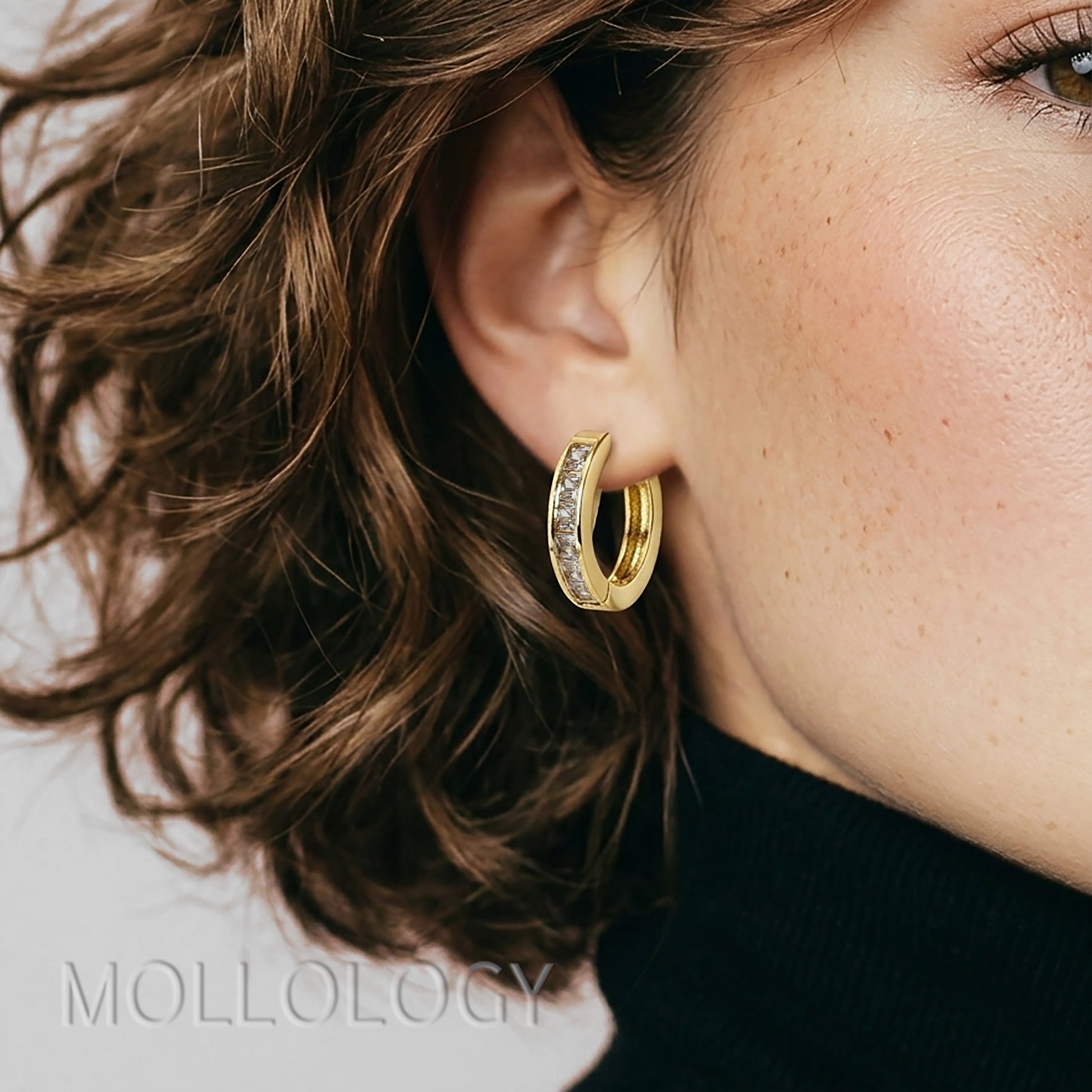 Close-up of a woman wearing gold hoop earrings with 'Mollogoy' branding.