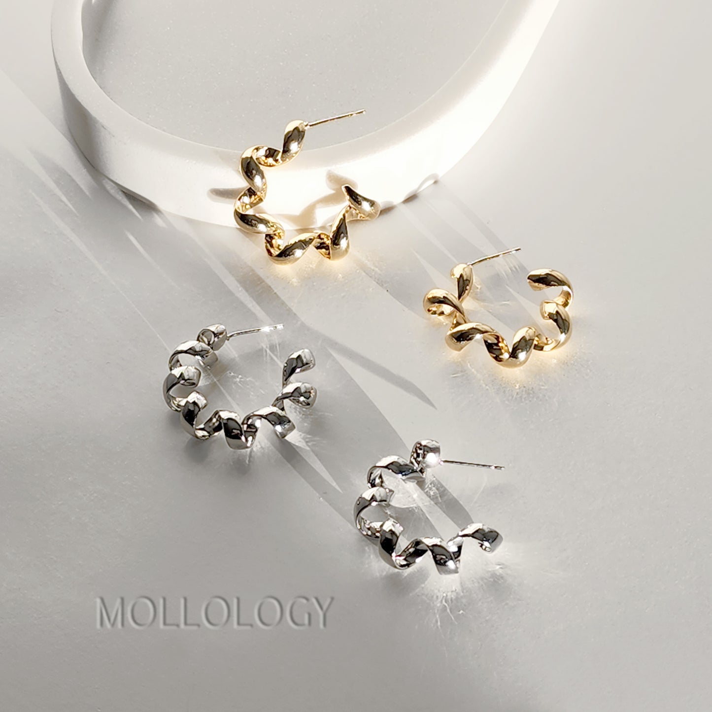 Three twisted hoop earrings displayed on a glossy white surface, including two gold-toned hoops and one silver-toned hoops, with the brand name MOLLOLOGY printed at the bottom left.