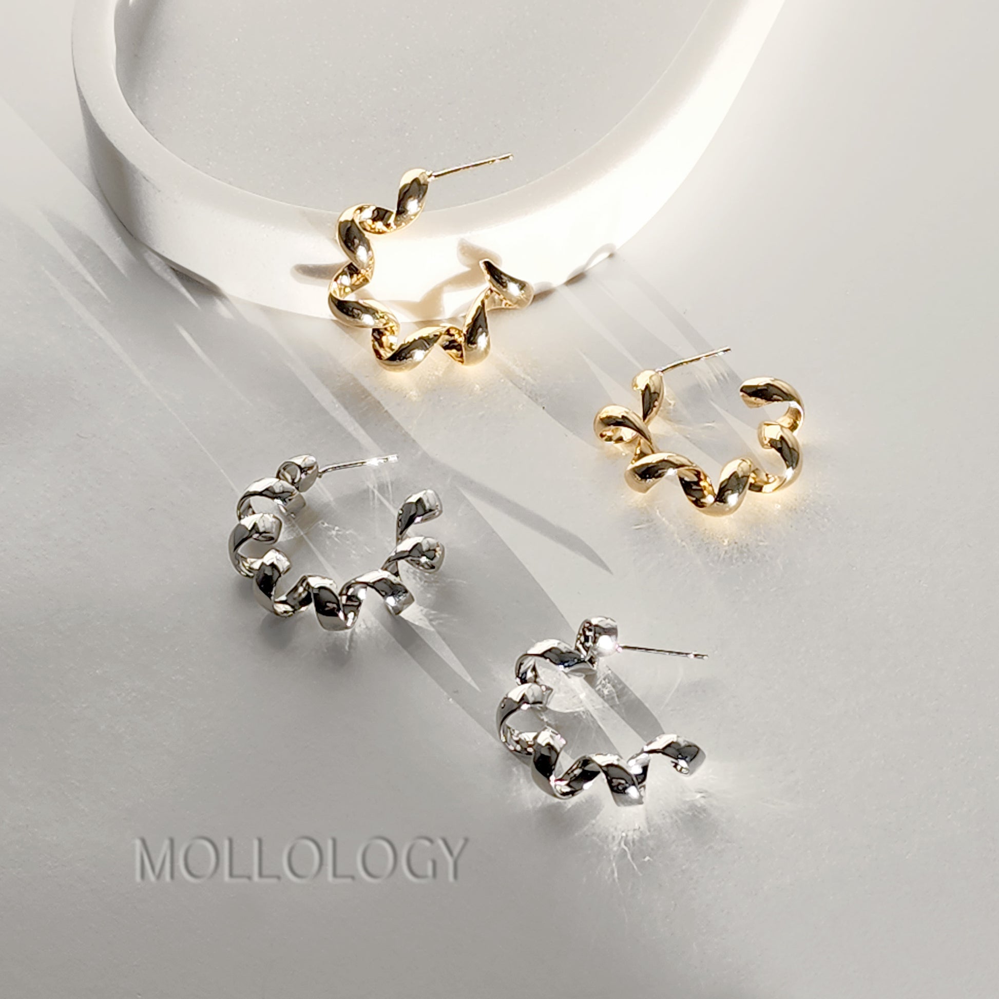 Three twisted hoop earrings displayed on a glossy white surface, including two gold-toned hoops and one silver-toned hoops, with the brand name MOLLOLOGY printed at the bottom left.