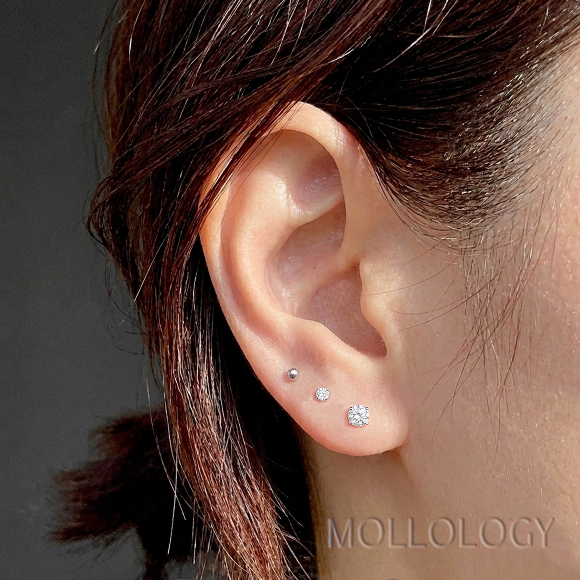 Close-up of a person's ear showing three small stud earrings in different sizes, with dark brown hair and a gray background. The word MOLLOLOGY appears at the bottom right.