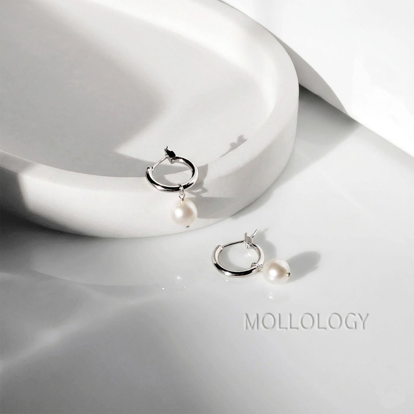 A pair of silver hoop earrings with a single white pearl drop, displayed on a glossy white surface next to a white ceramic dish. The brand name MOLLOLOGY is printed on the surface.