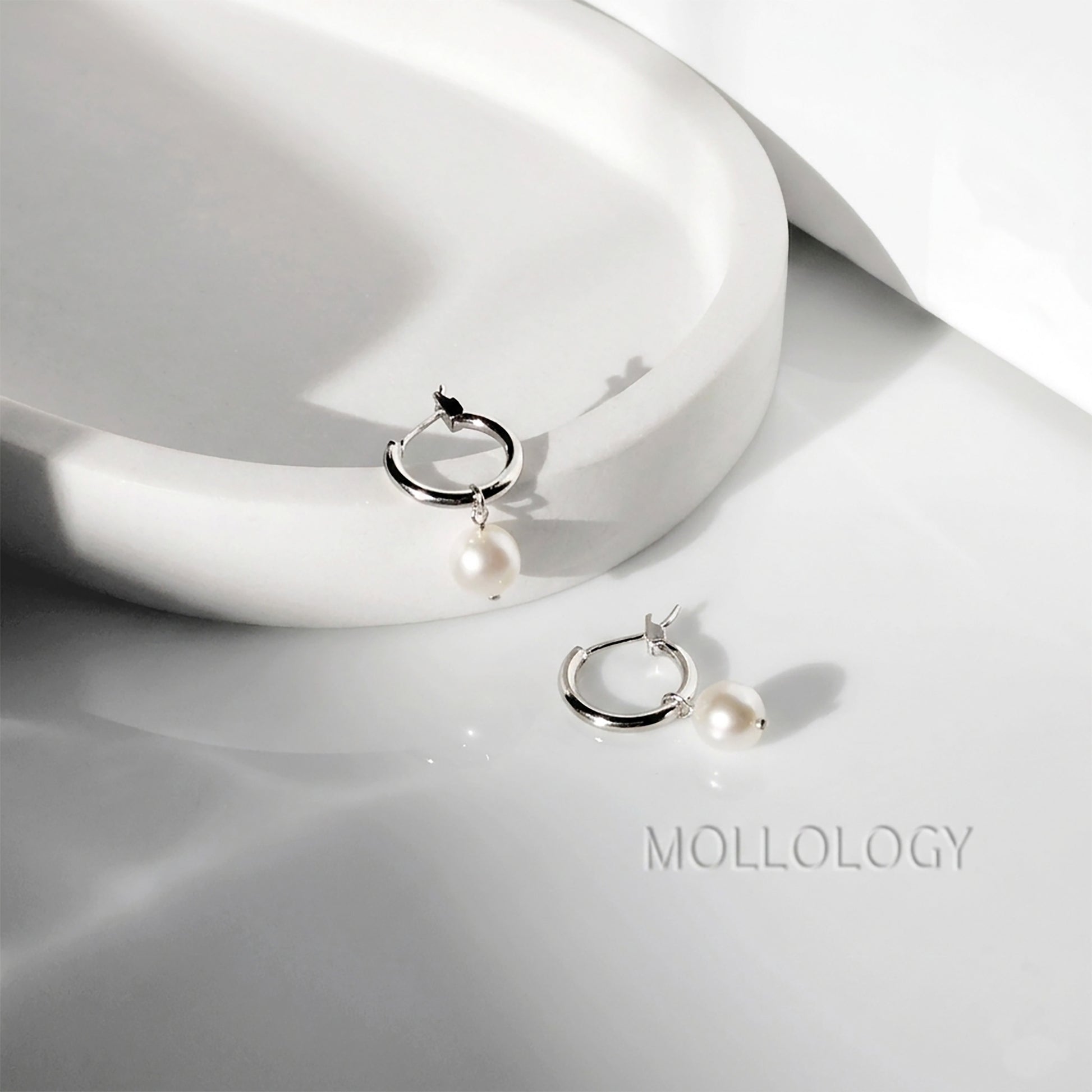 A pair of silver hoop earrings with a single white pearl drop, displayed on a glossy white surface next to a white ceramic dish. The brand name MOLLOLOGY is printed on the surface.