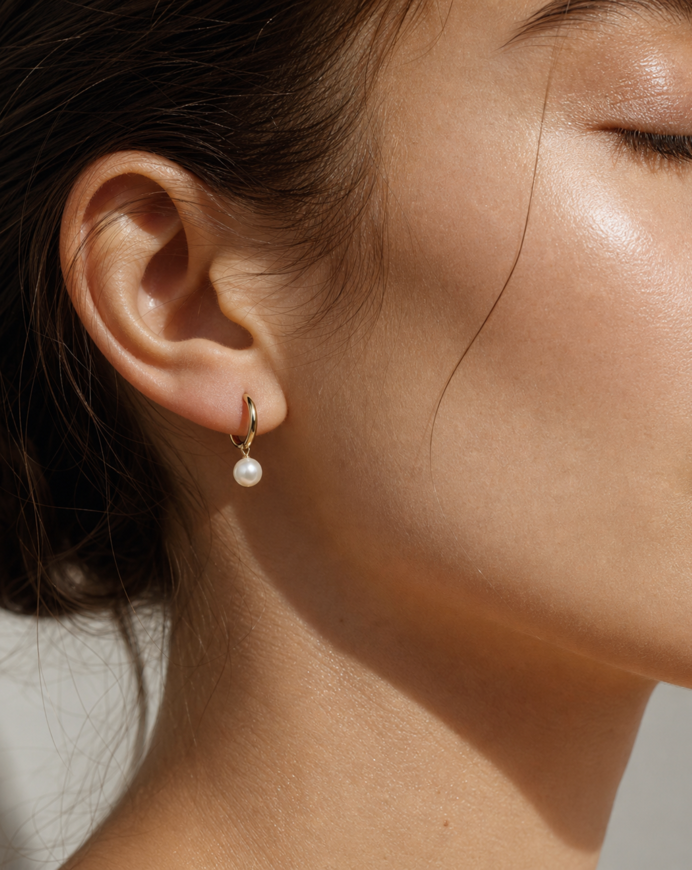 A close-up of a model's ear wearing a small gold huggie hoop earring with a dangling white pearl.
