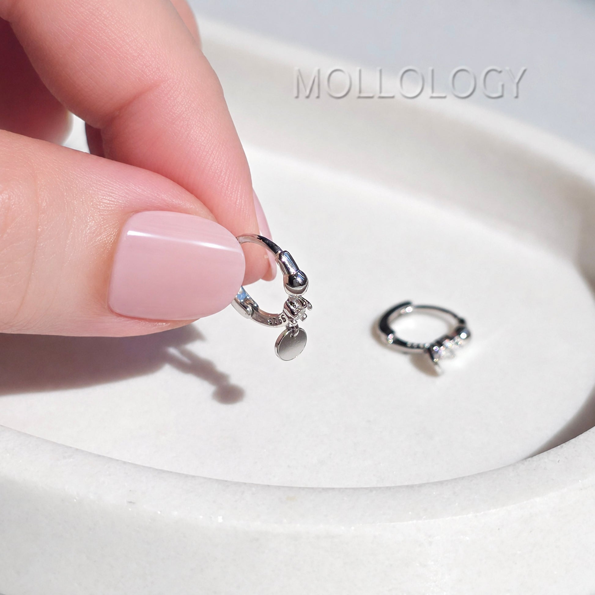 A close-up of a hand with pale pink manicured nails holding a small silver huggie hoop earring with a dangling charm, while another matching silver hoop lies on a white ceramic dish. The brand name MOLLOLOGY is faintly visible in the upper right.
