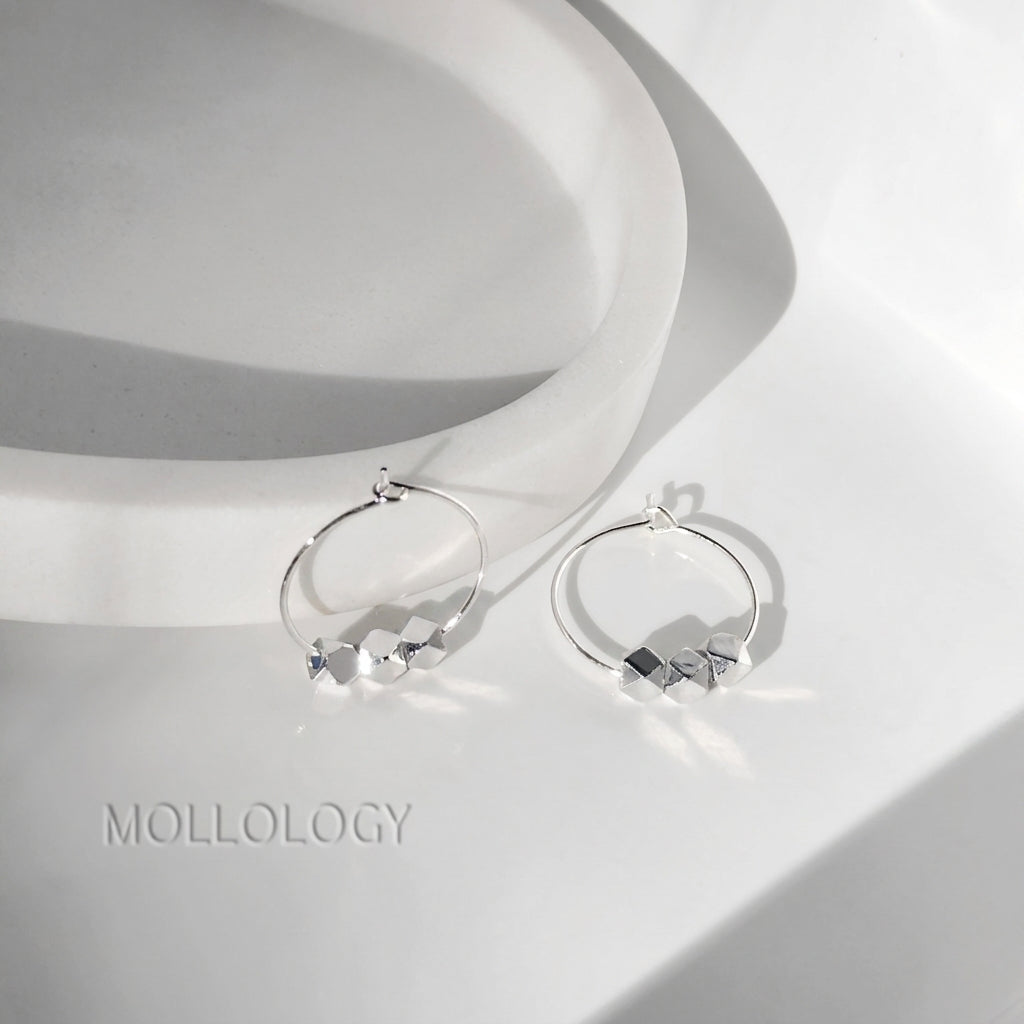 A pair of silver hoop earrings with small geometric diamond-shaped charms, displayed on a white ceramic pedestal with a soft shadow. The brand name 'MOLLOLOGY' is printed in gray text in the lower-left corner.