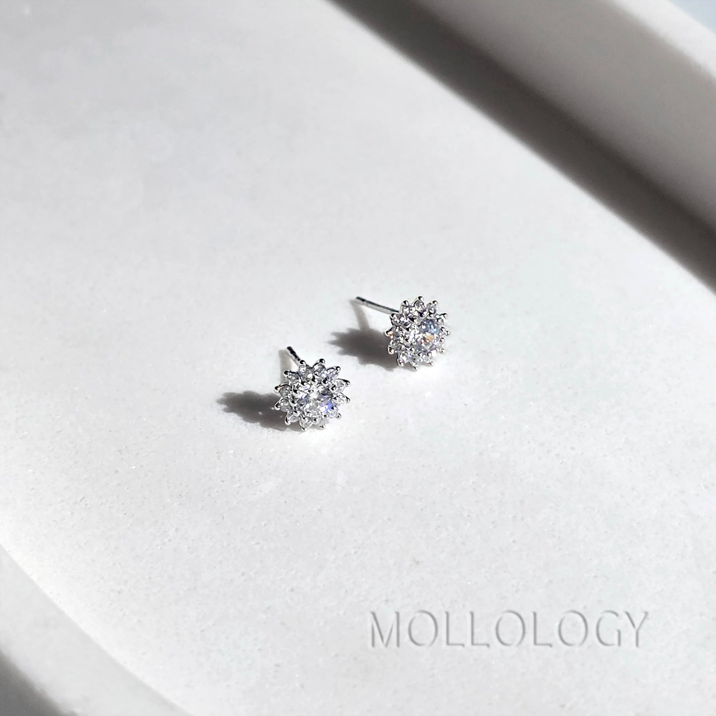 A pair of small silver stud earrings with clear gemstones, displayed on a white textured surface with a soft shadow. The brand name MOLLOLOGY is printed in the lower right corner.