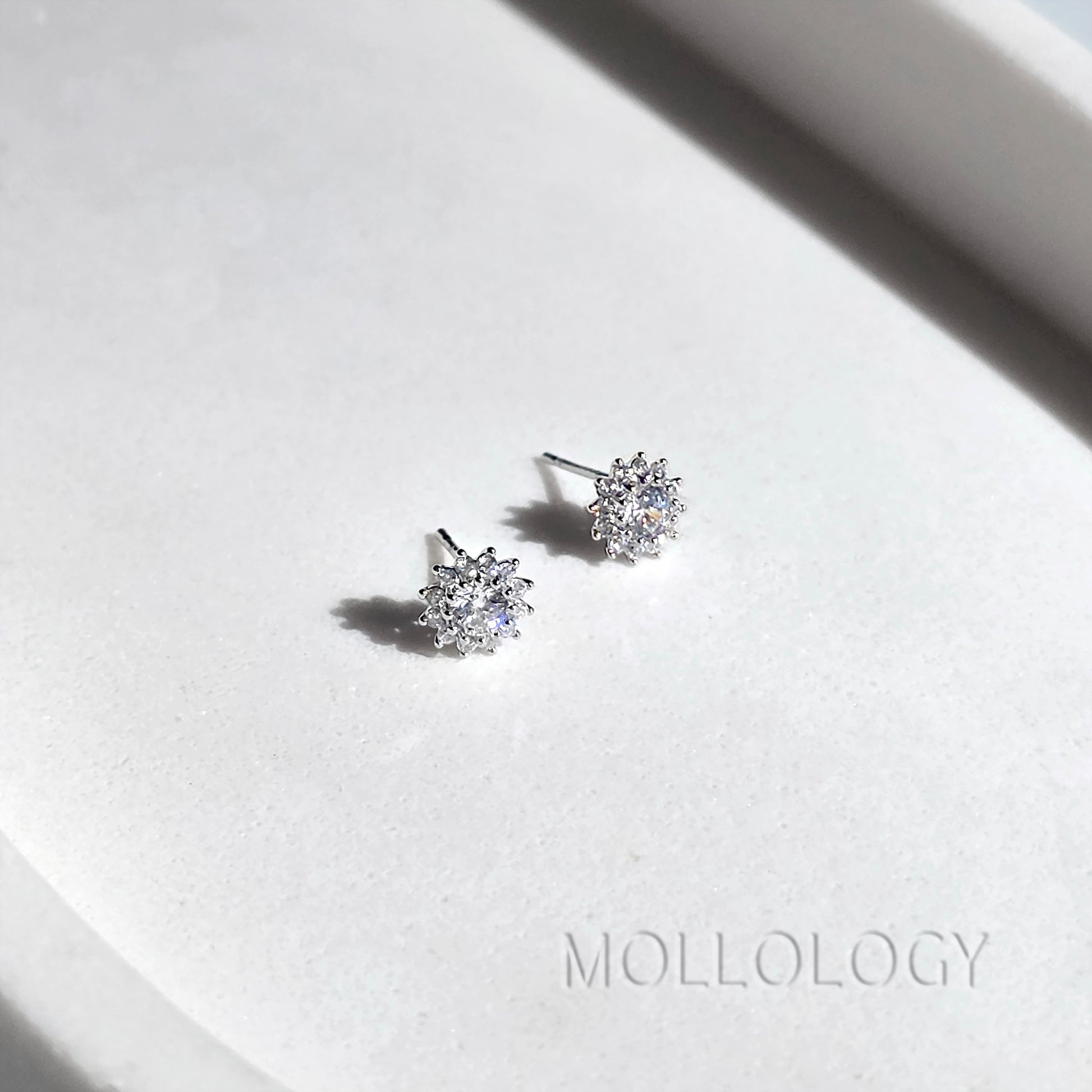 A pair of small silver stud earrings with clear gemstones, displayed on a white textured surface with a soft shadow. The brand name MOLLOLOGY is printed in the lower right corner.
