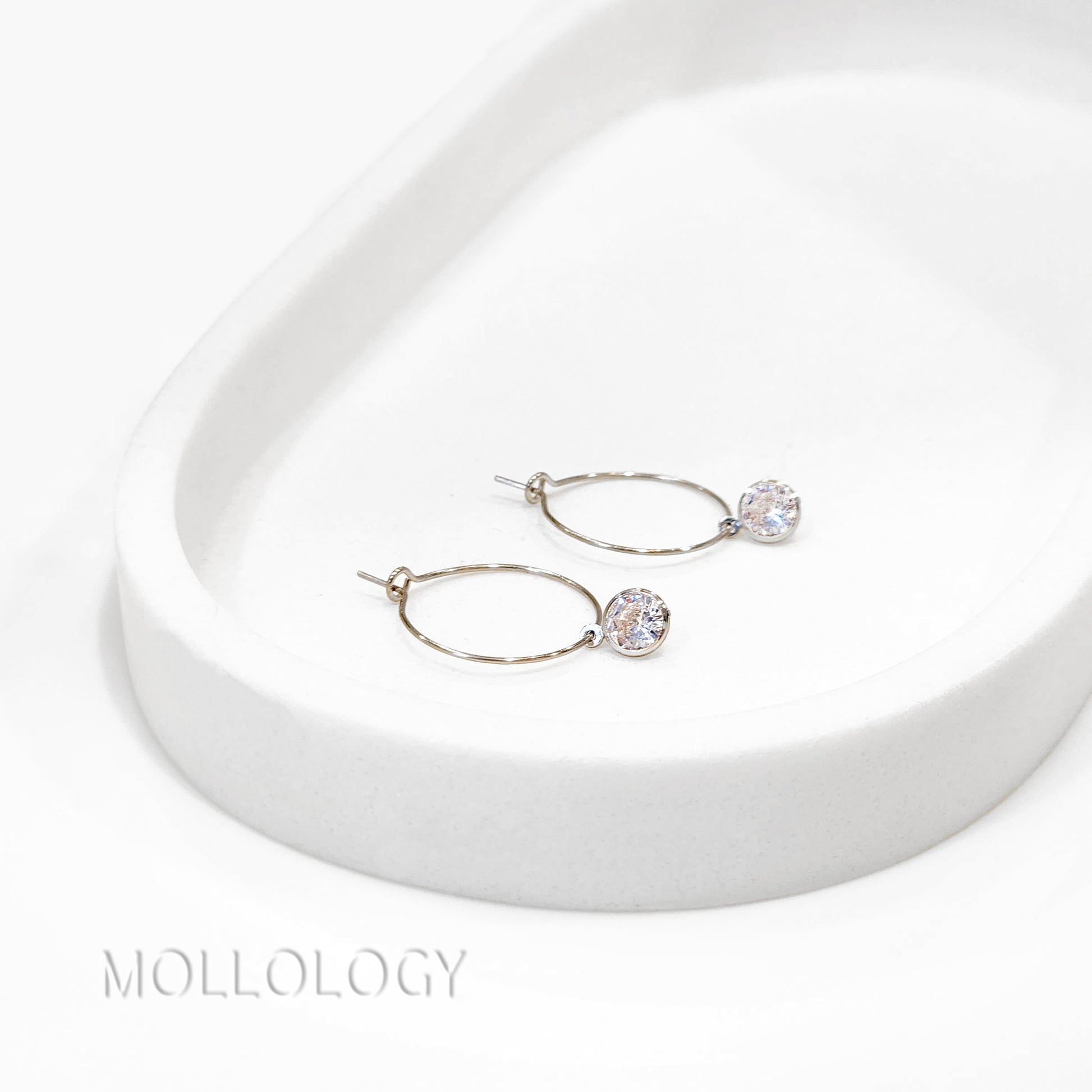 Pair of silver hoop earrings with clear gemstones on a white surface, branded 'Molloy'.