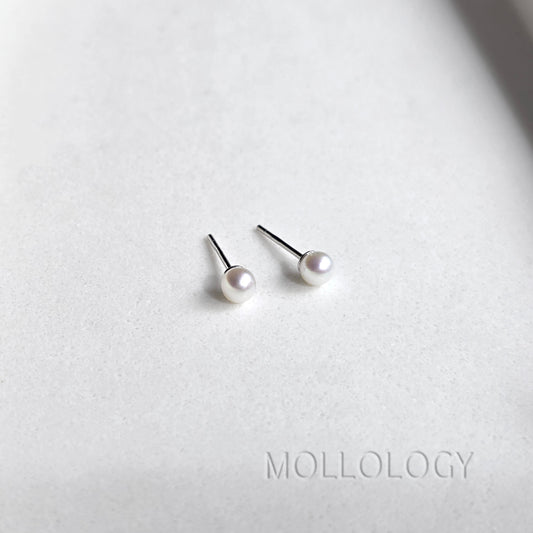 A pair of minimalist stud earrings featuring small white pearls on silver posts, displayed on a textured light gray surface with the brand name MOLLOLOGY printed at the bottom.