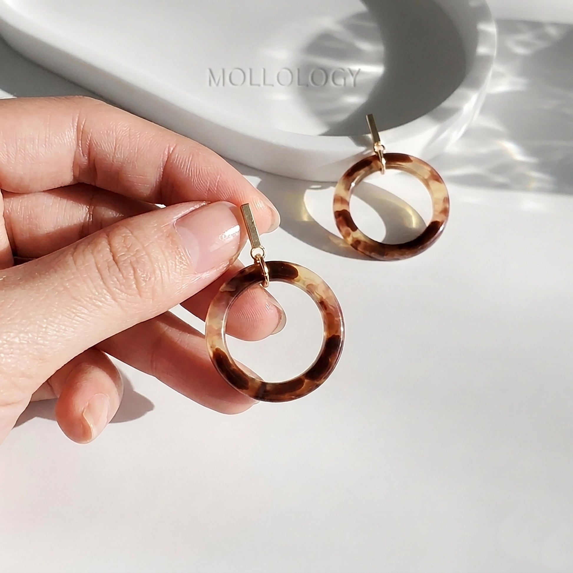 Close-up of hand holding tortoiseshell resin hoop drop earrings showing gold rectangular bar stud post and brown marbled acrylic ring pendant