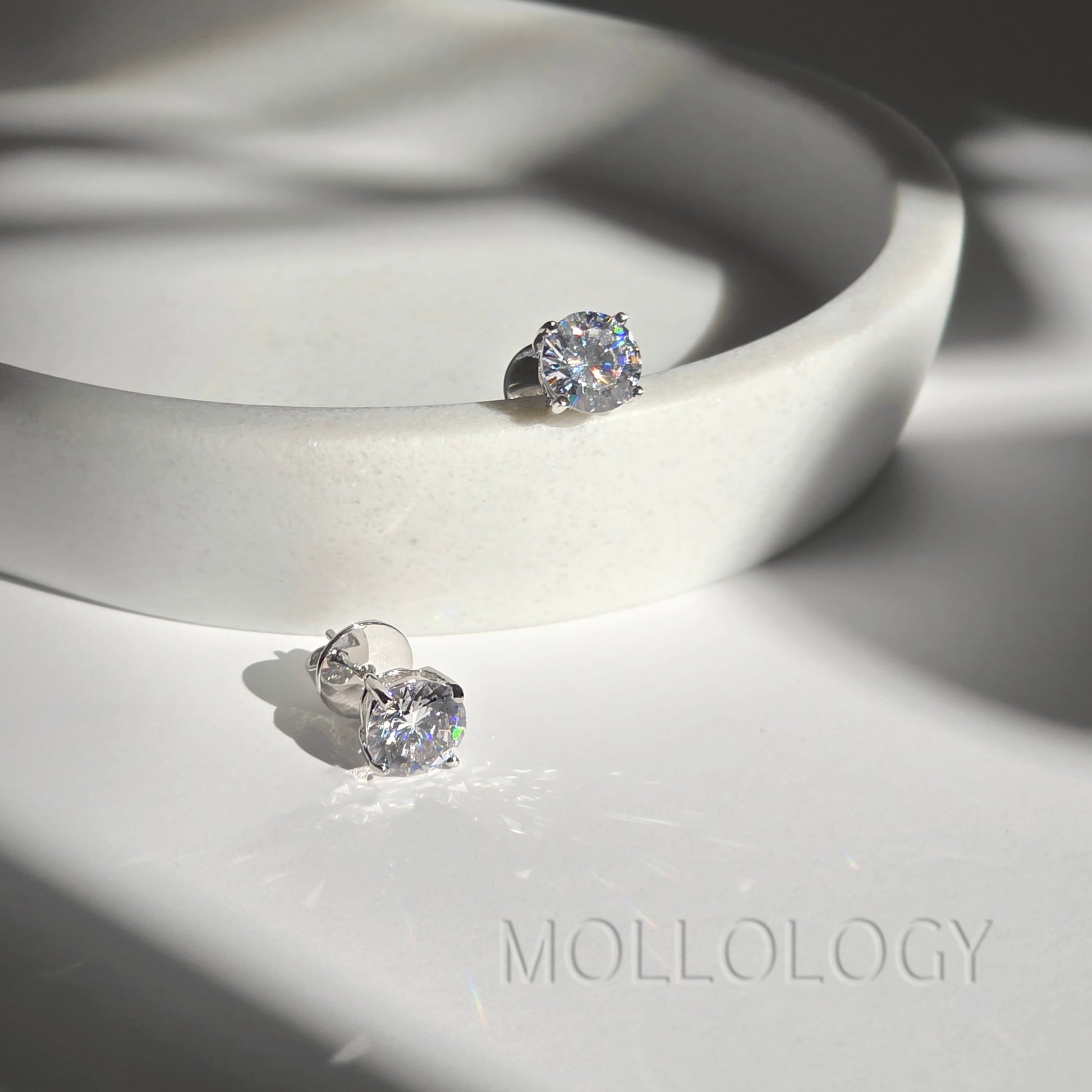 Pair of white gold round CZ stud earrings on curved white dish, minimalist diamond-style studs with shadows