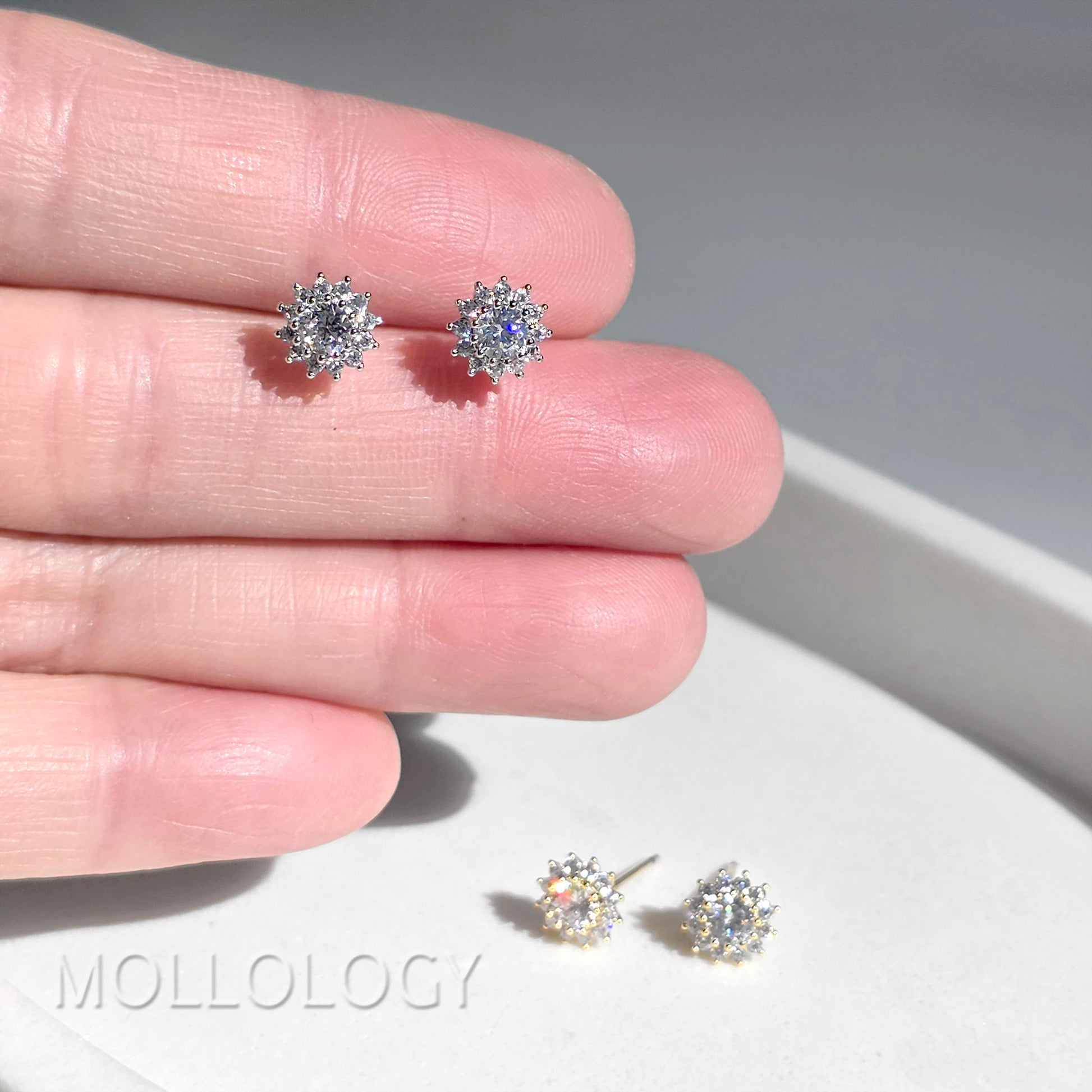 A close-up of a person's fingers holding a pair of small round stud earrings, with two matching earrings resting on a white surface below. The earrings appear silver-toned with clear stones.