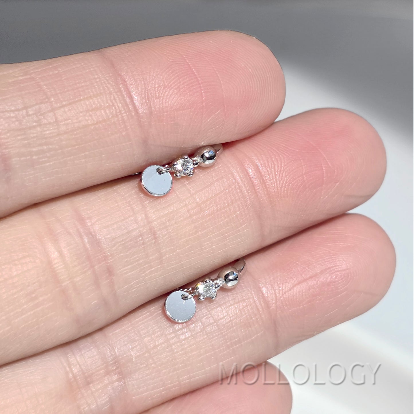 A close-up of a hand displaying a pair of small silver earrings with a flat circular disc charm, a clear faceted stone, and a polished bead, with the brand name MOLLOLOGY visible as a watermark.