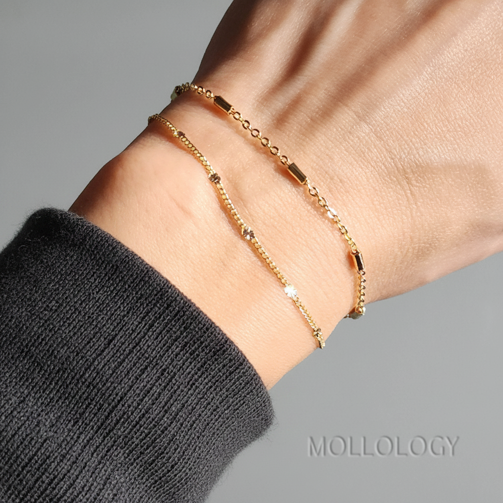 Two delicate gold chain bracelets with small rectangular and circular links, shown worn on a person's wrist. The person is wearing a dark gray ribbed sweater sleeve, against a neutral gray background with the brand name MOLLOLOGY in the bottom right corner.
