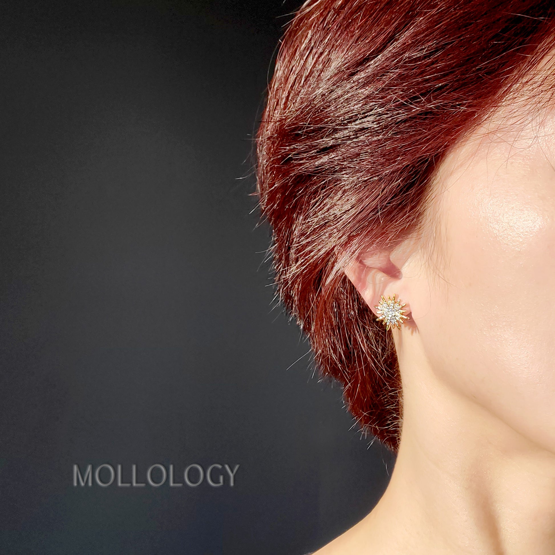 A close-up side profile of a person with short reddish-brown hair wearing a small gold-toned stud earring with clear stones, against a dark gray background. The word MOLLOLOGY appears in the lower left.