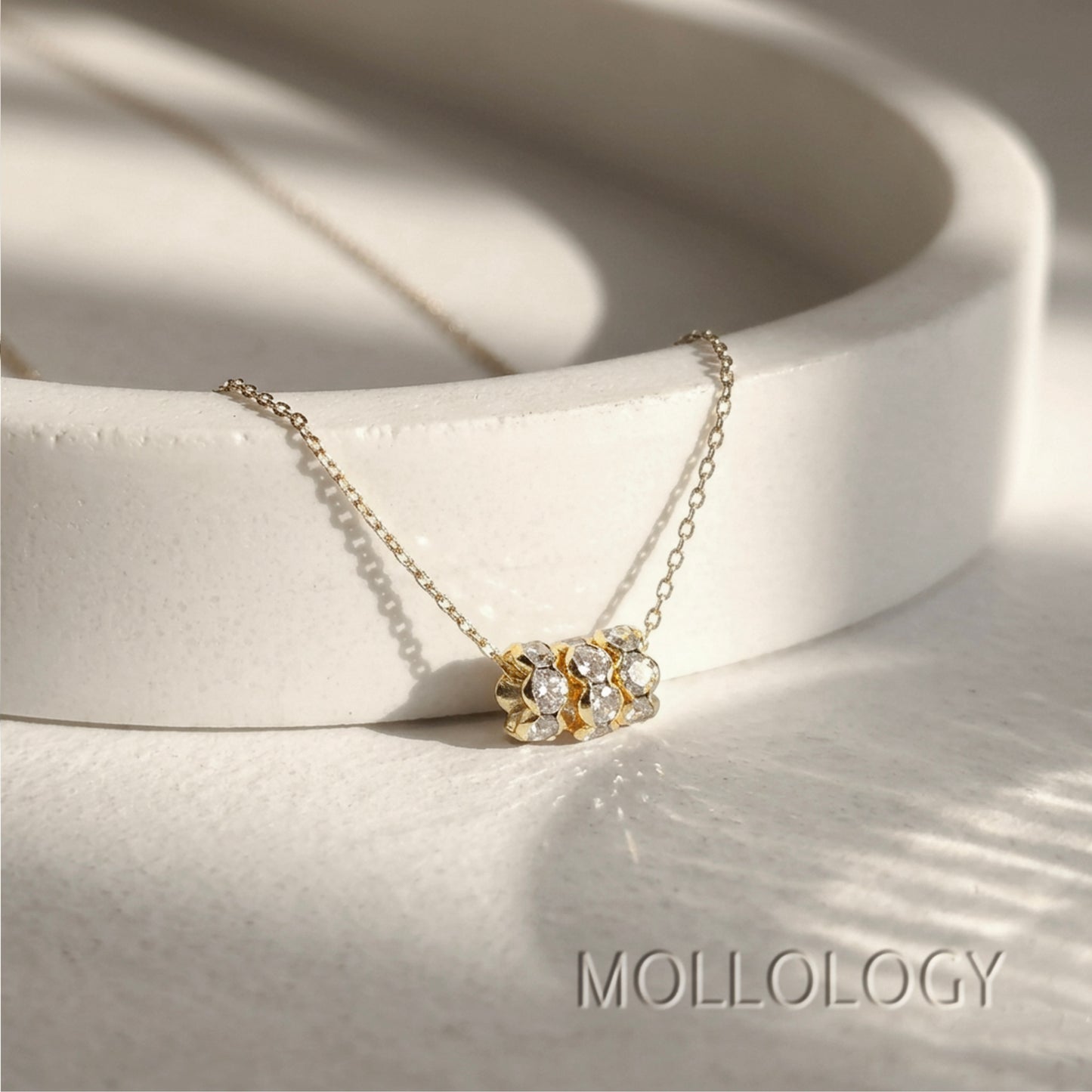 A delicate gold chain necklace with a small cluster of clear gemstones in a gold setting, displayed on a white ceramic dish. The brand name 'MOLLOLOGY' is printed in gray text in the bottom right corner.