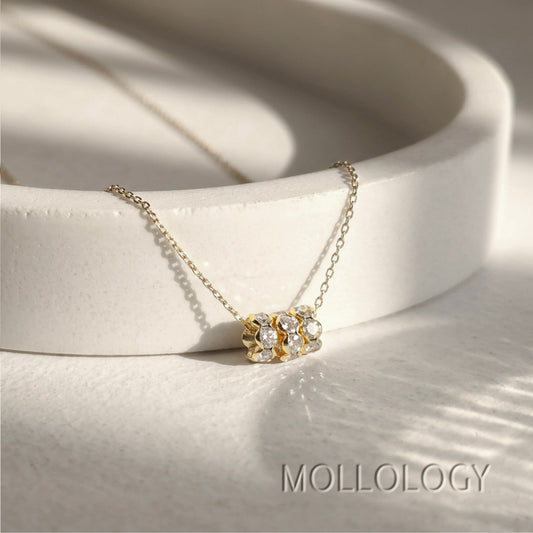 A delicate gold chain necklace with a small cluster of clear gemstones in a gold setting, displayed on a white ceramic dish. The brand name 'MOLLOLOGY' is printed in gray text in the bottom right corner.