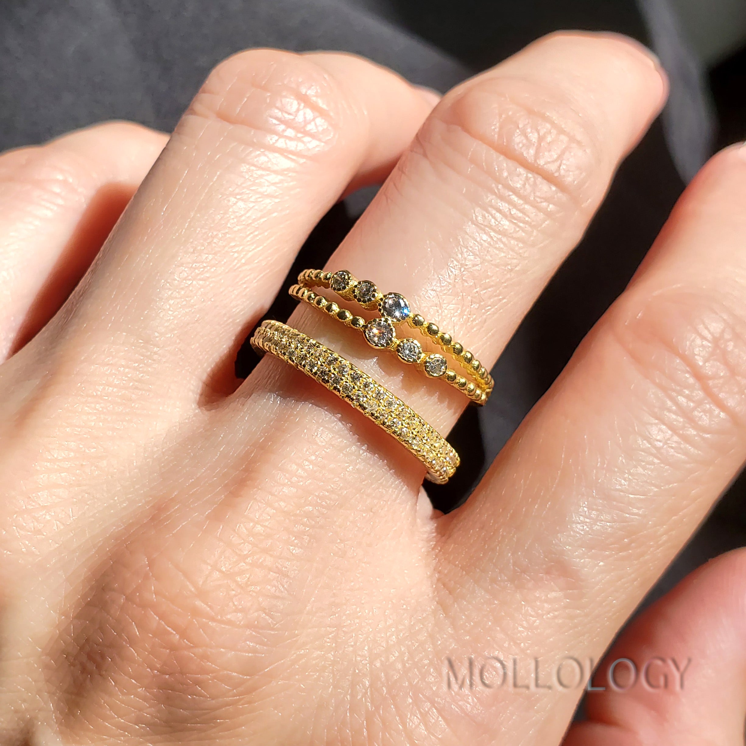 Close-up of a hand wearing two gold rings with gemstones on a neutral background.