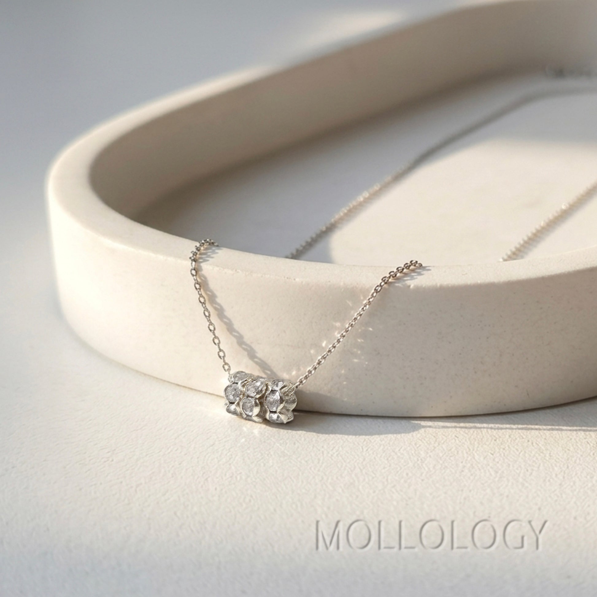 A delicate silver necklace with a small cluster of clear gemstones on a fine chain, draped over a curved cream-colored ceramic tray. The brand name MOLLOLOGY is printed in a subtle gray font in the bottom right corner.