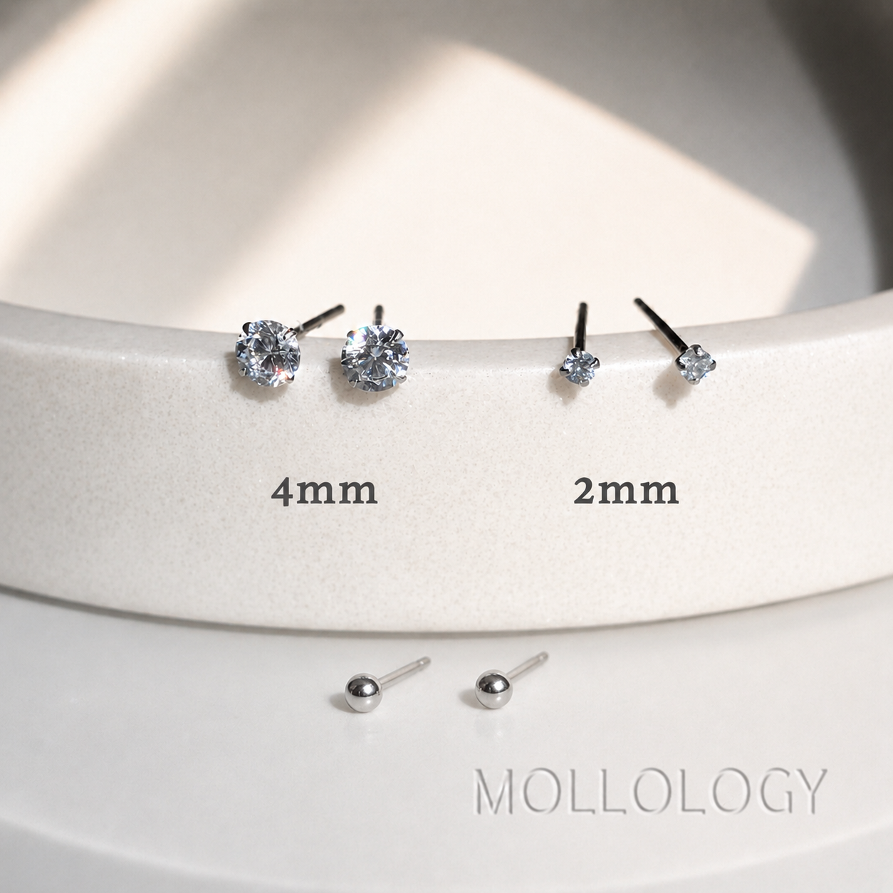 Two pairs of silver stud earrings shown on a white display stand, one labeled 4mm and one labeled 2mm, with two loose studs below. The brand name MOLLOLOGY appears at the bottom right.