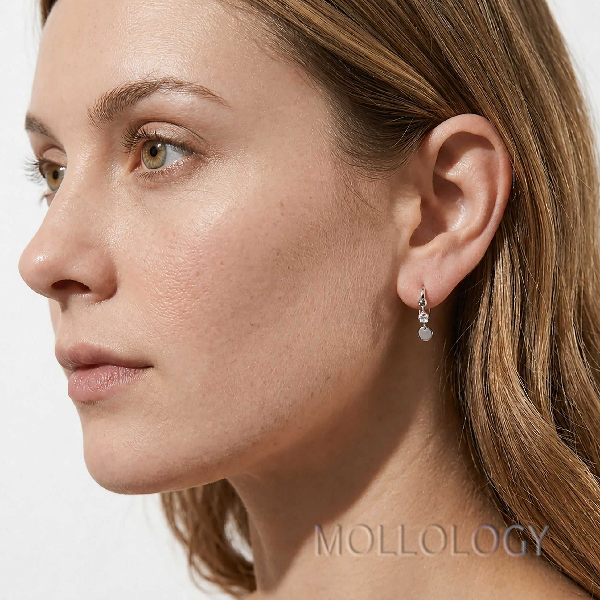 Close-up side profile of a woman wearing a delicate silver-toned dangle earring with a small clear stone, against a light neutral background. The brand name MOLLOLOGY is visible as a watermark at the bottom right.