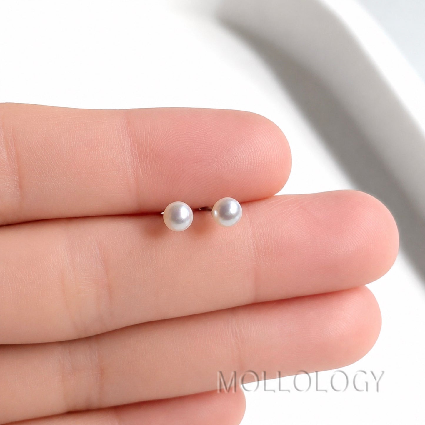 A pair of small pearl stud earrings held between fingers for scale, with a white background and the MOLLOLOGY brand name visible at the bottom.