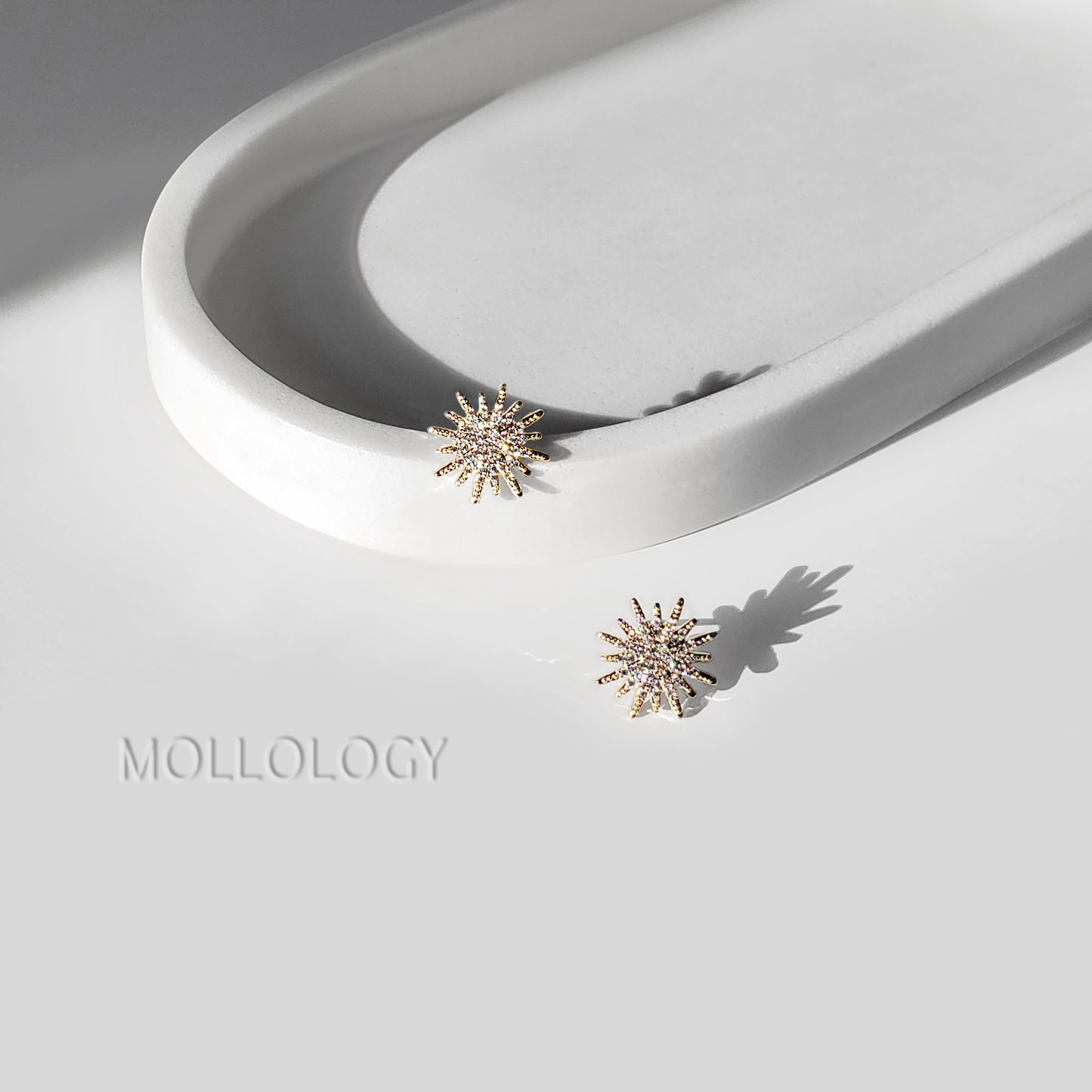Two gold-toned starburst stud earrings with small clear stones, displayed on a white sculptural tray and white surface with the MOLLOLOGY brand name.