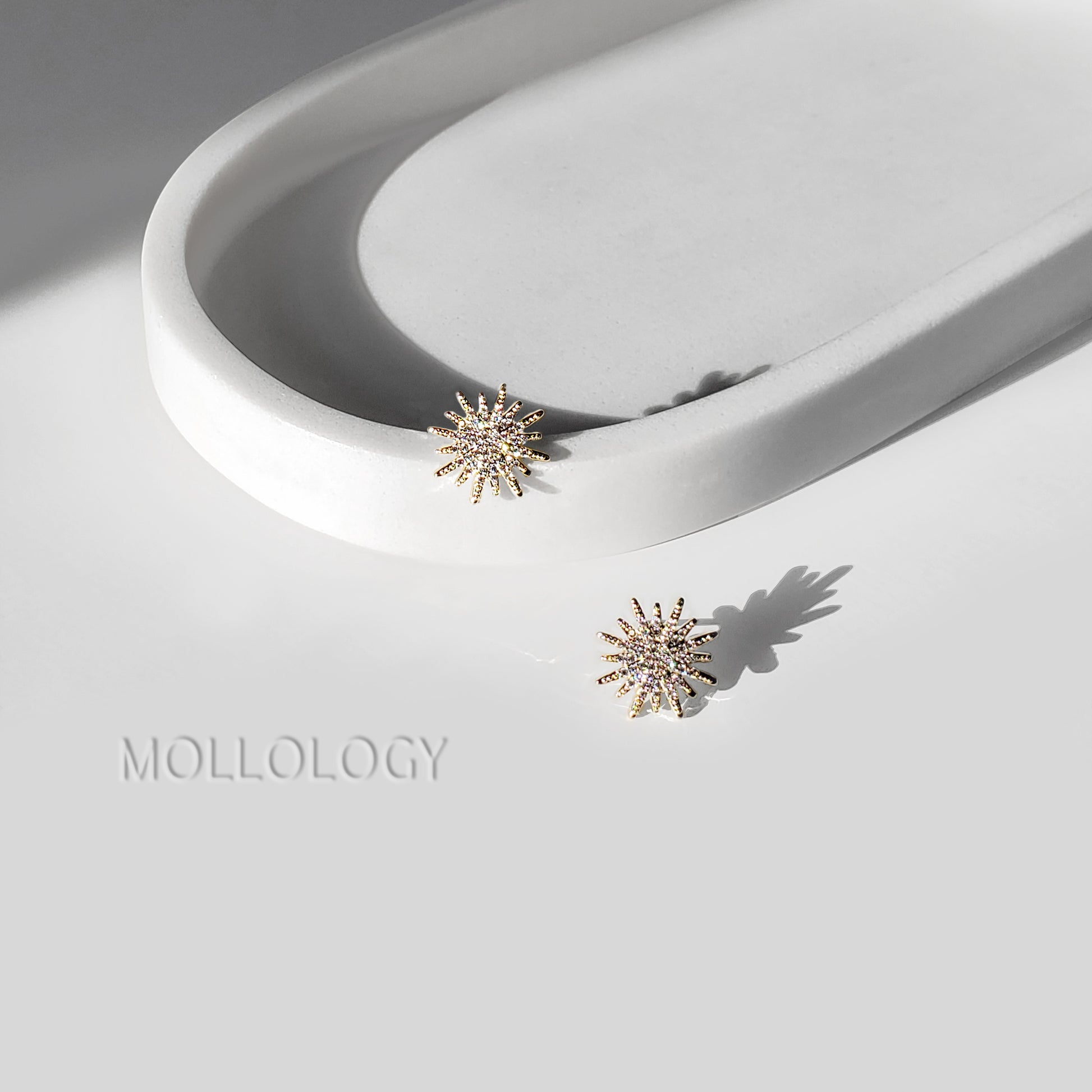 Two gold-toned starburst stud earrings with small clear stones, displayed on a white sculptural tray and white surface with the MOLLOLOGY brand name.