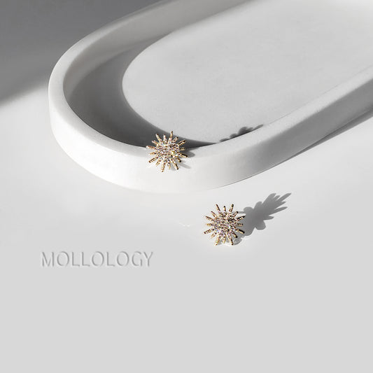 Two gold-toned starburst stud earrings with small clear stones, displayed on a white sculptural tray and white surface with the MOLLOLOGY brand name.