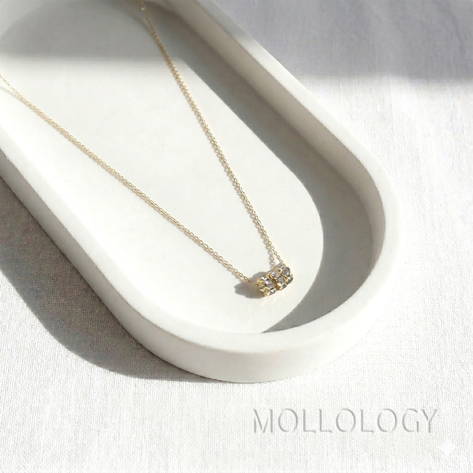 A delicate gold chain necklace with a small box pendant set with clear crystals, displayed on a white ceramic tray. The brand name 'MOLLOLOGY' is printed in a subtle font in the bottom right corner.