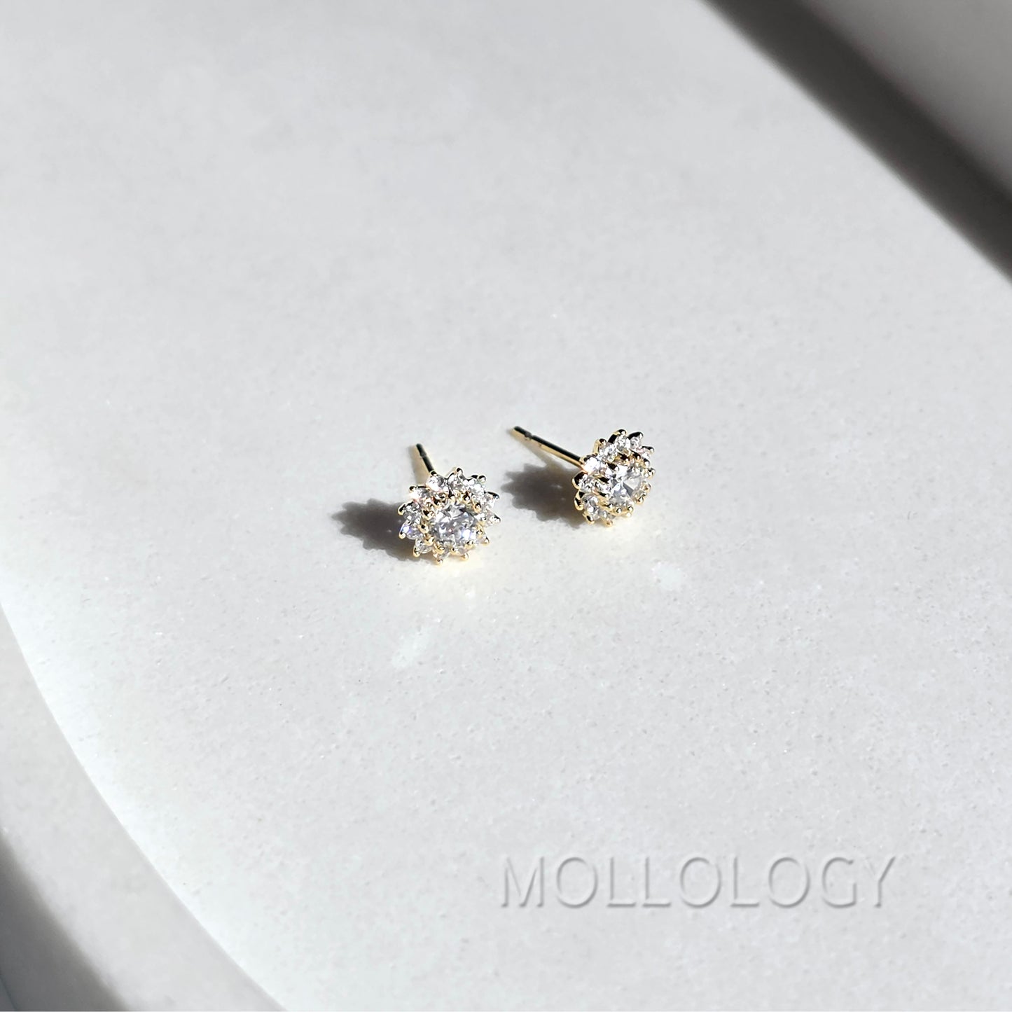 A pair of gold stud earrings with clear gemstones, displayed on a white textured surface with a soft shadow. The brand name 'MOLLOLOGY' is faintly visible in the bottom right corner.