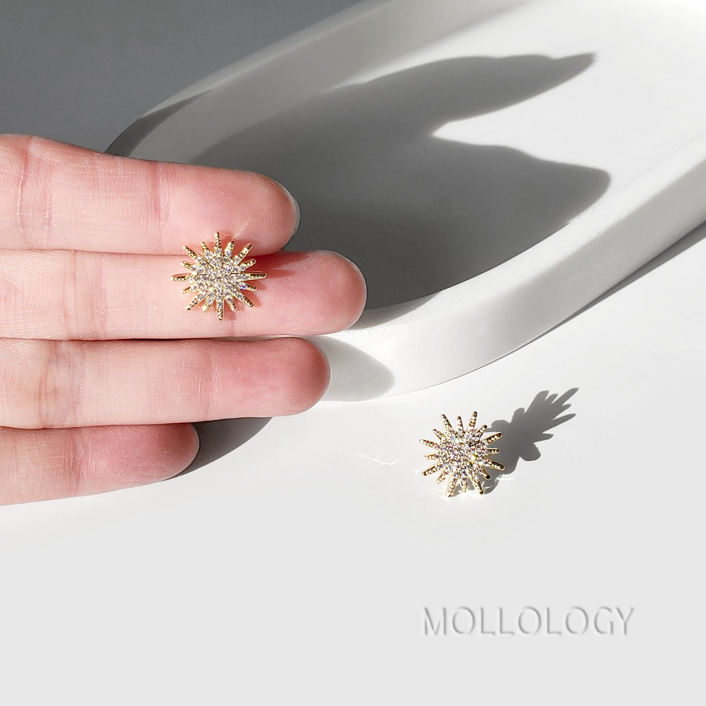 A hand holds a pair of gold-toned sunburst stud earrings with clear stones, with one earring resting on a white surface below. The brand name MOLLOLOGY is printed in the lower right corner.