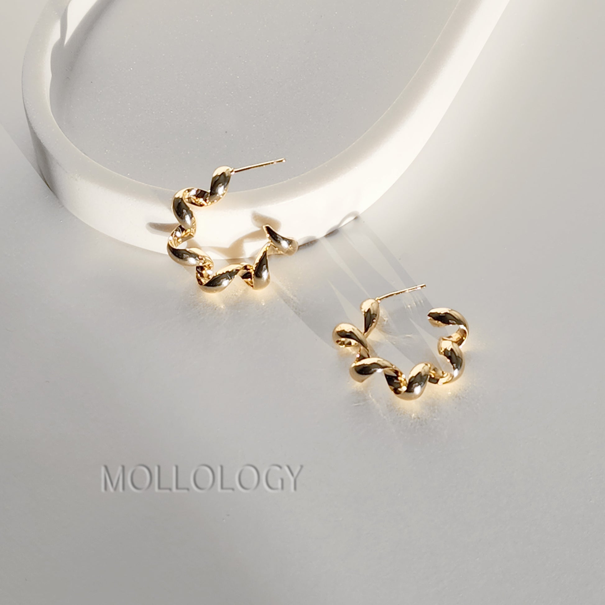 A pair of gold huggie hoop earrings with a twisted, leaf-like organic design, displayed on a light gray surface next to a white curved prop. The brand name 'MOLLOLOGY' is printed on the surface below.