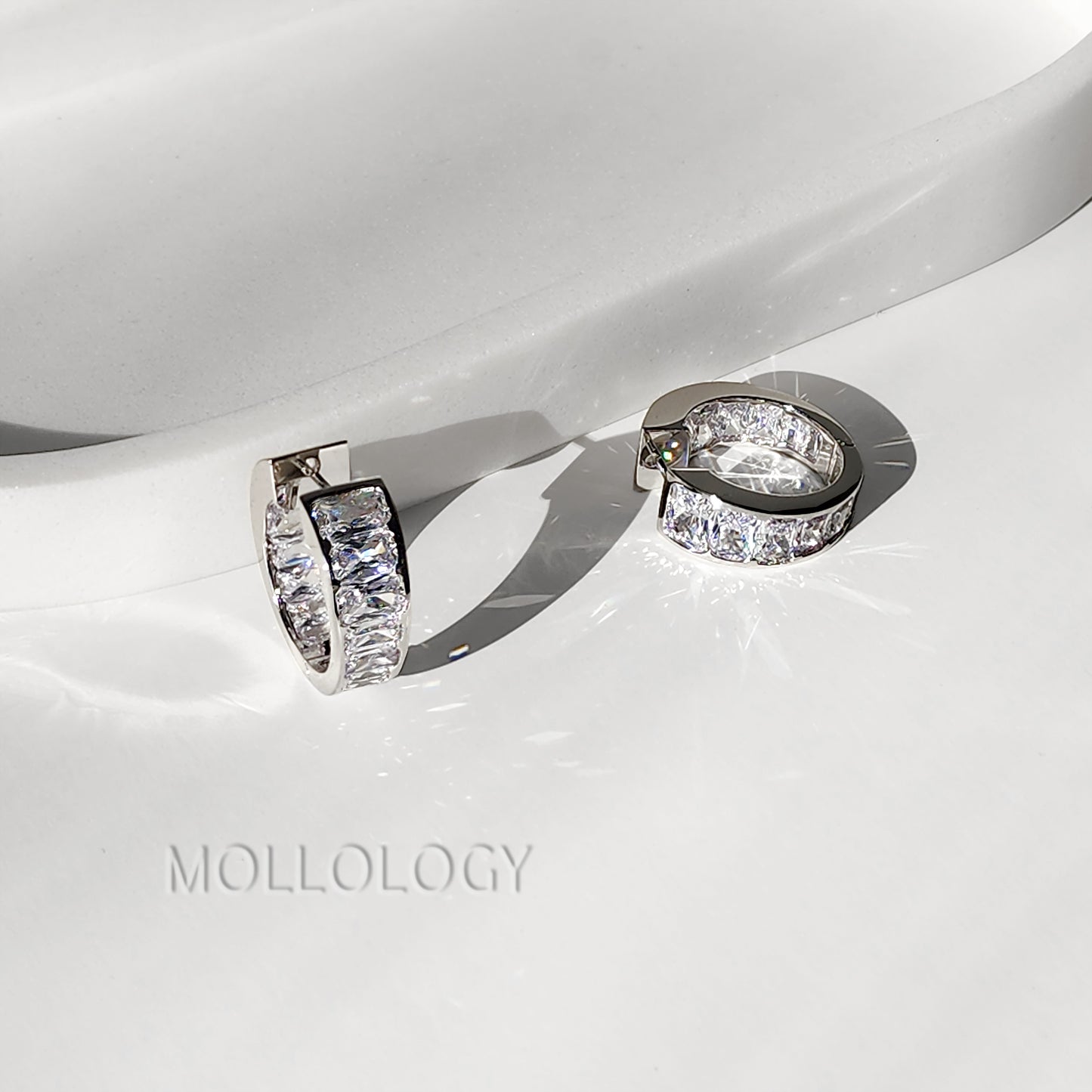 A pair of silver-toned huggie hoop earrings with clear rectangular pavé stones, displayed on a glossy white surface with a soft shadow. The brand name 'MOLLOLOGY' is faintly visible in the lower left.