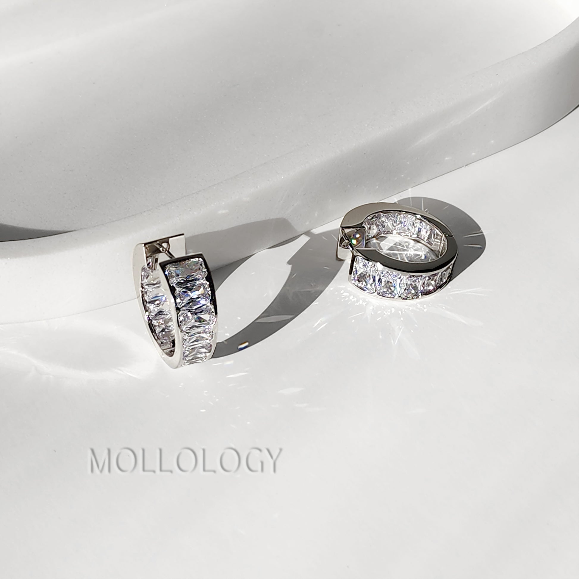 A pair of silver-toned huggie hoop earrings with clear rectangular pavé stones, displayed on a glossy white surface with a soft shadow. The brand name 'MOLLOLOGY' is faintly visible in the lower left.