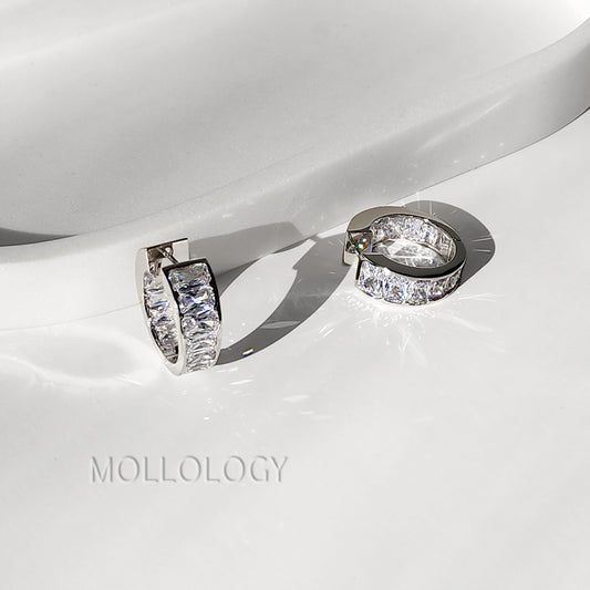 A pair of silver-toned huggie hoop earrings with clear rectangular pavé stones, displayed on a glossy white surface with a soft shadow. The brand name 'MOLLOLOGY' is faintly visible in the lower left.