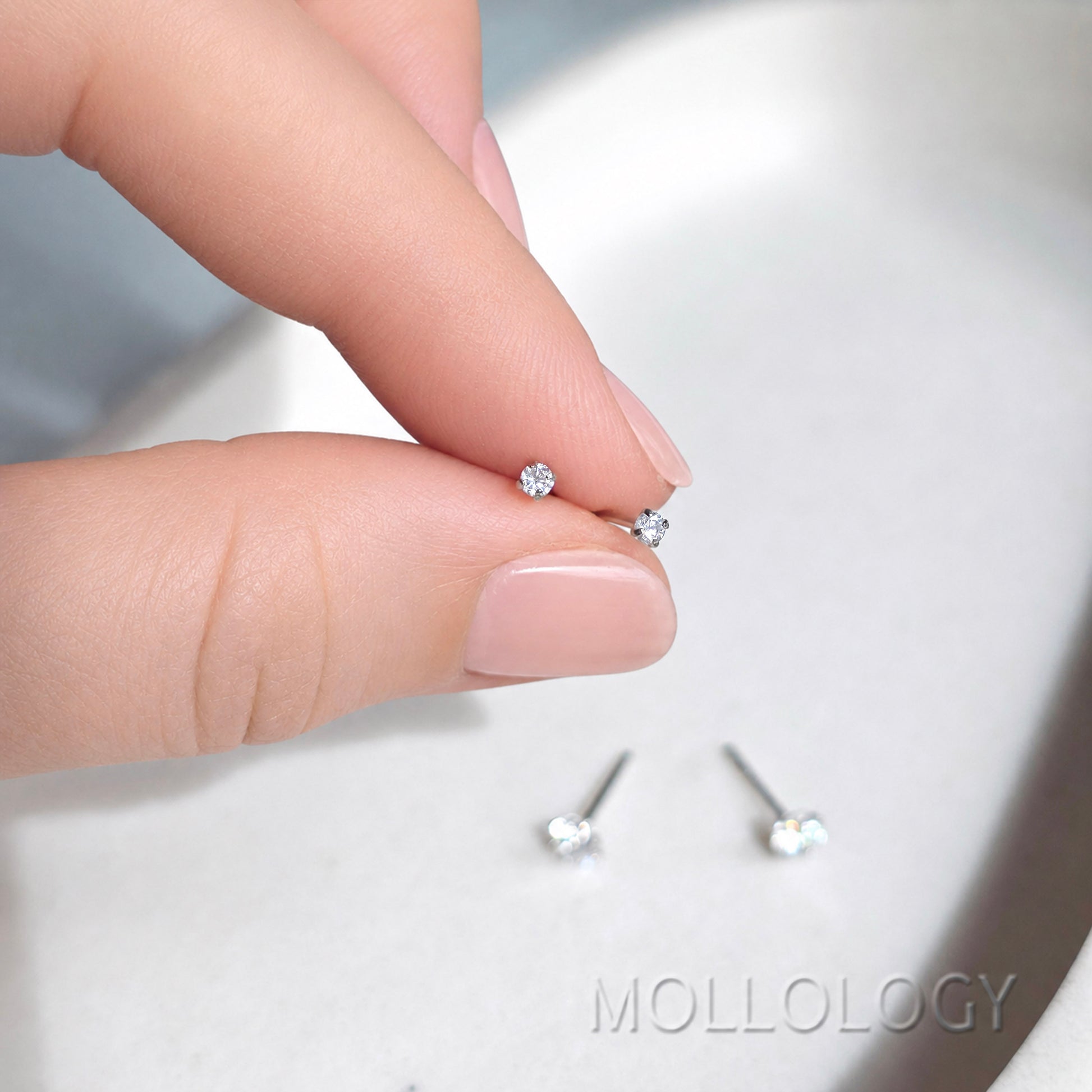 A close-up of a hand holding two tiny silver stud earrings with clear gemstones, with a pair lying below on a light surface. The brand name MOLLOLOGY is visible in the bottom right corner.