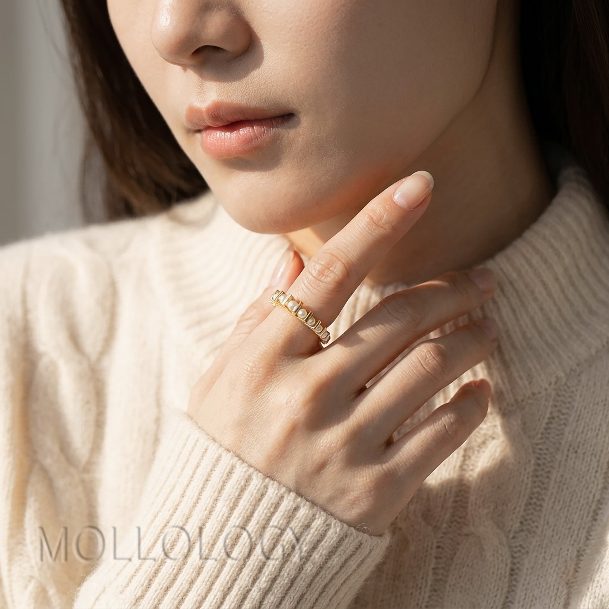 Model in cream sweater wearing dainty gold pearl ring near chin, cropped closeup portrait, minimalist stacking pearl jewelry with nude manicure
