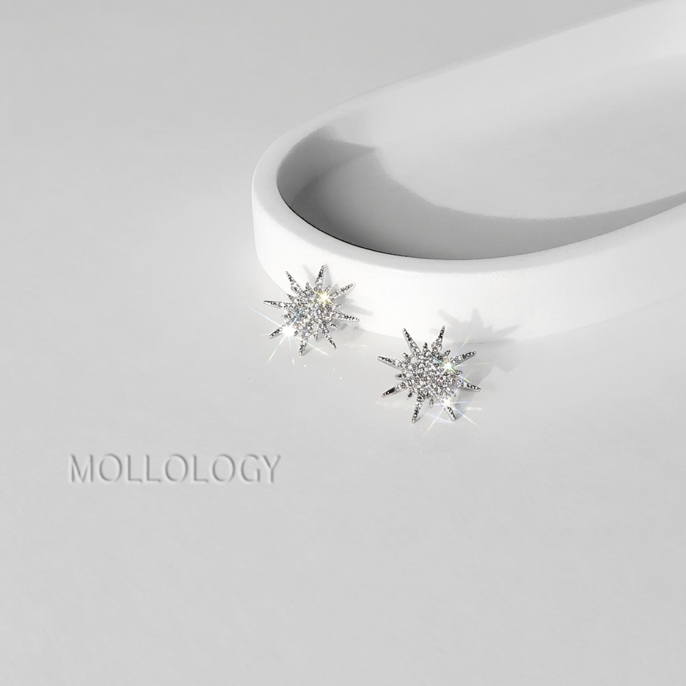 A pair of silver-toned starburst stud earrings with clear sparkling stones, displayed on a white curved stand against a light gray background. The brand name MOLLOLOGY is printed in a subtle gray font on the lower left.