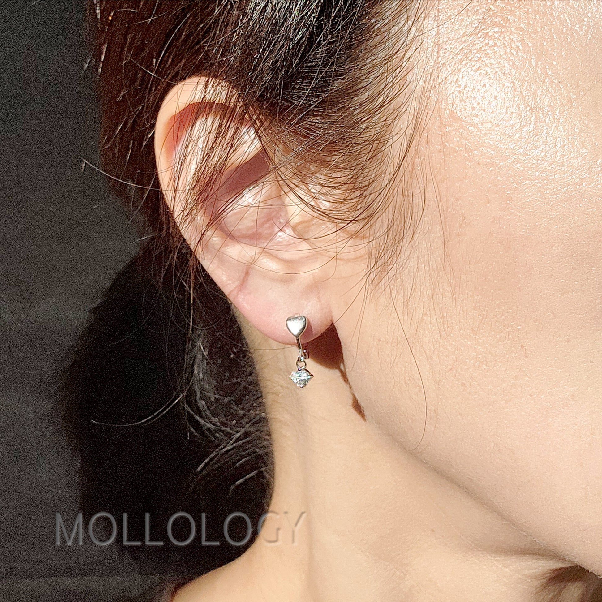 Close-up of an ear wearing a heart-shaped earring with a small dangling gemstone, branded 'Mollology'.