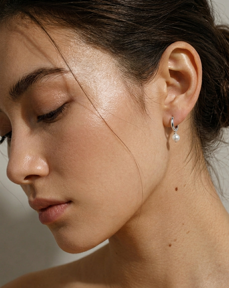 A close-up of a woman's ear wearing a small silver hoop earring with a dangling white pearl, shown against a soft neutral background.