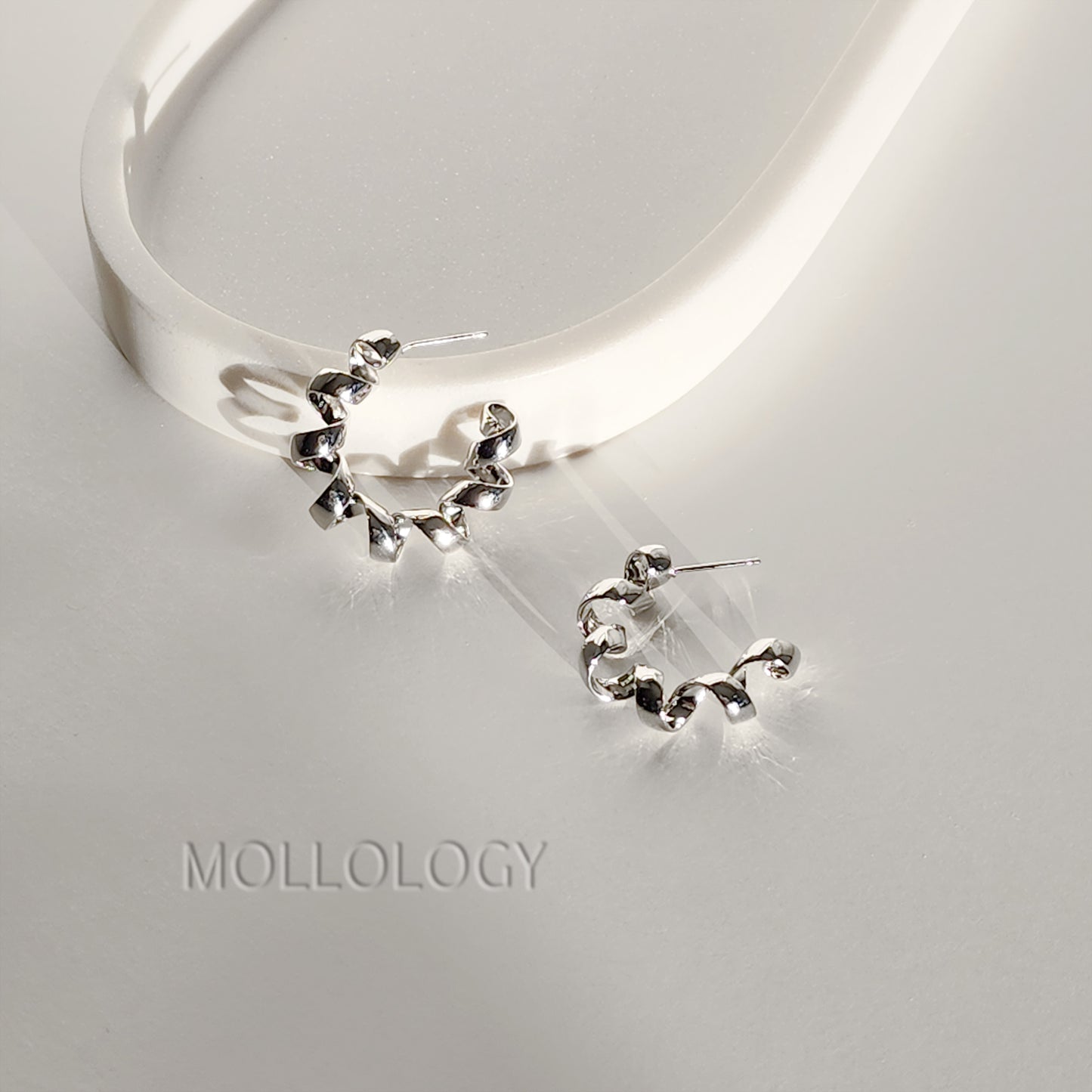 A pair of silver crescent-shaped hoop earrings with a textured, hammered finish, displayed on a light gray surface next to a white curved display stand. The brand name 'MOLLOLOGY' is subtly printed on the surface below.